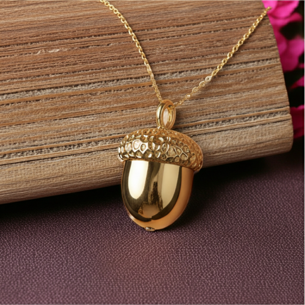 The Gold Acorn Pendant Necklace in 24K gold plate by MokshaGoldSculptures, shown on rustic wood with pink blooms.